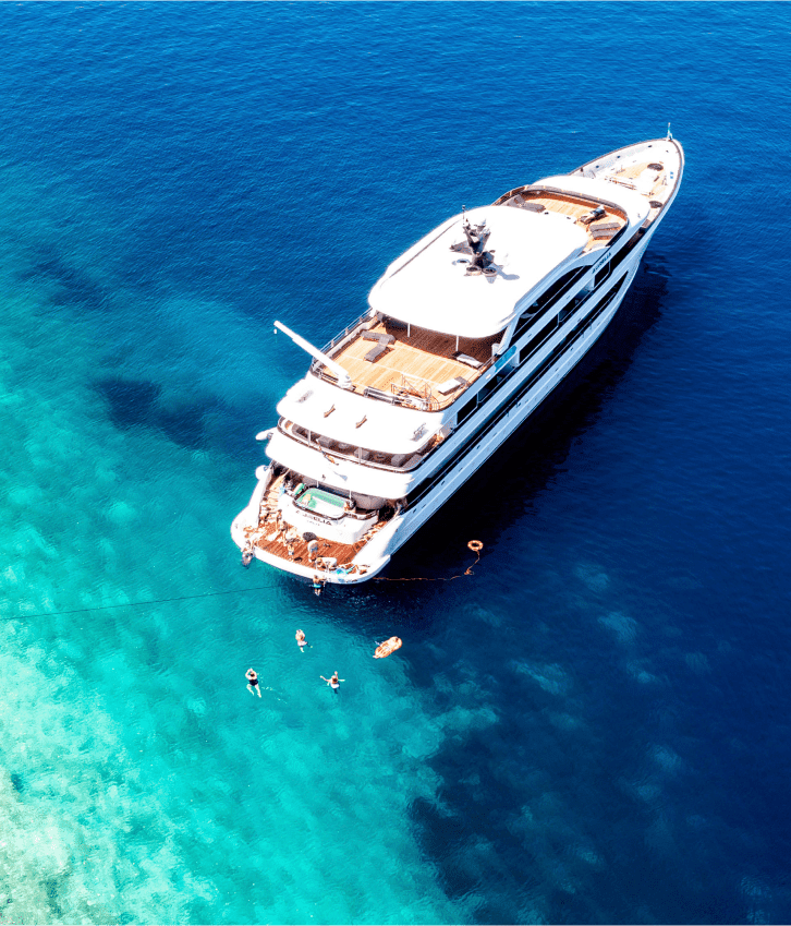 An aerial view of a multi-deck luxury motor yacht anchored in crystal-clear turquoise Mediterranean