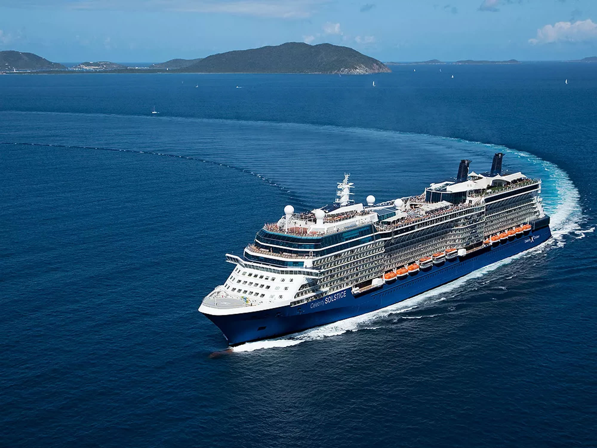 A large modern cruise ship with distinctive blue hull and orange lifeboats cruises through deep blue waters near a mountainous island coastline. Multiple sailboats are visible in the background, with lush green hills and clear skies completing this tropical seascape.