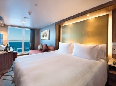 A spacious, modern cruise ship cabin featuring a king-sized bed with premium linens, an illuminated