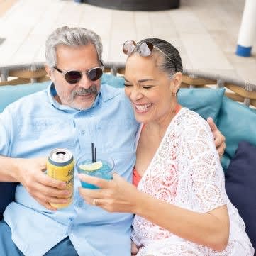 A middle-aged couple sits together on a cruise ship deck, smiling while holding tropical drinks
