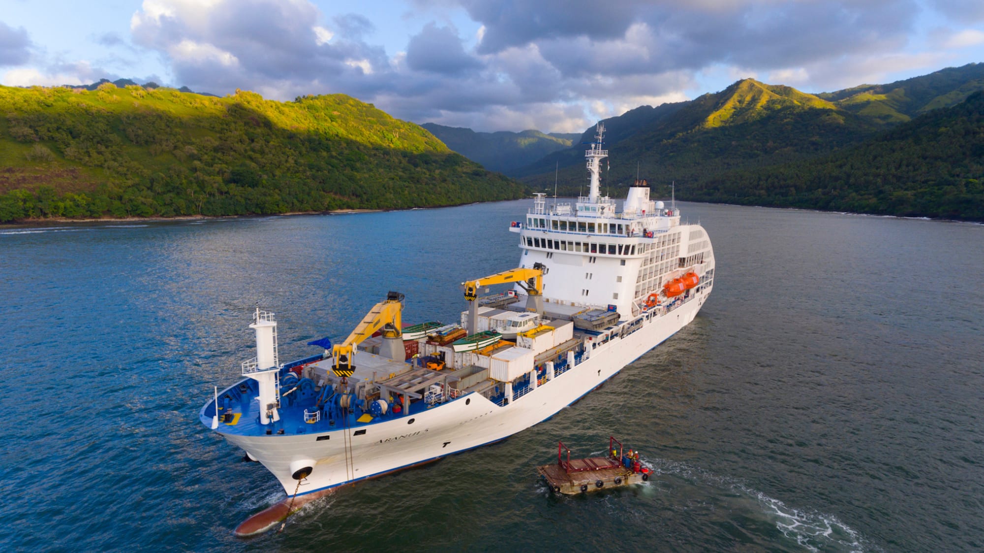 A white research or supply vessel navigating through a picturesque bay with lush green mountainous coastline, accompanied by a small support boat or platform.