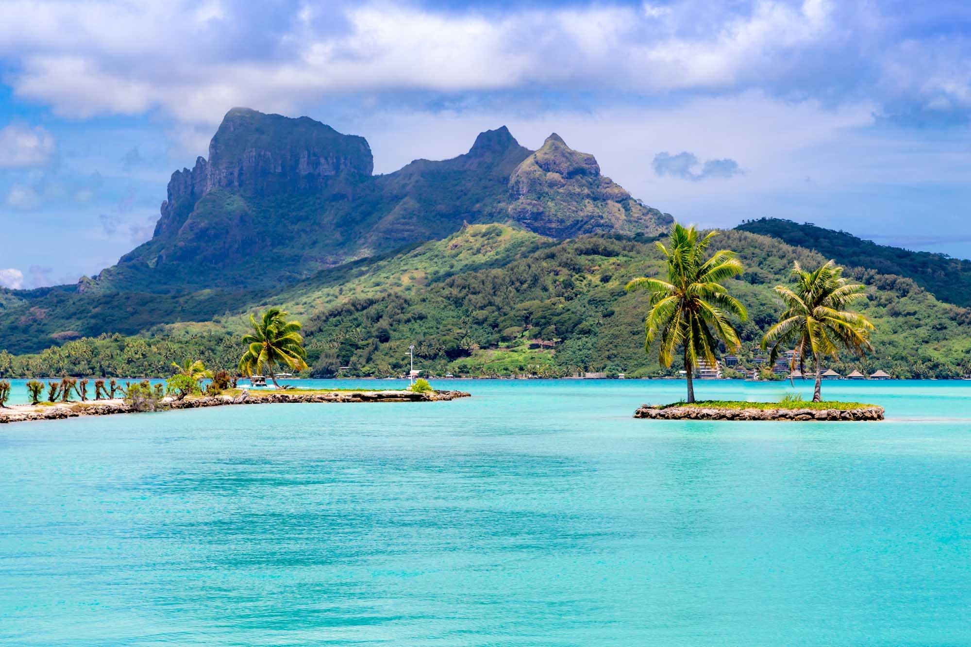 A stunning tropical landscape featuring the distinctive volcanic peaks of Bora Bora rising dramatically from lush green slopes, framed by crystal-clear turquoise lagoon waters dotted with coconut palms and small islets in French Polynesia.