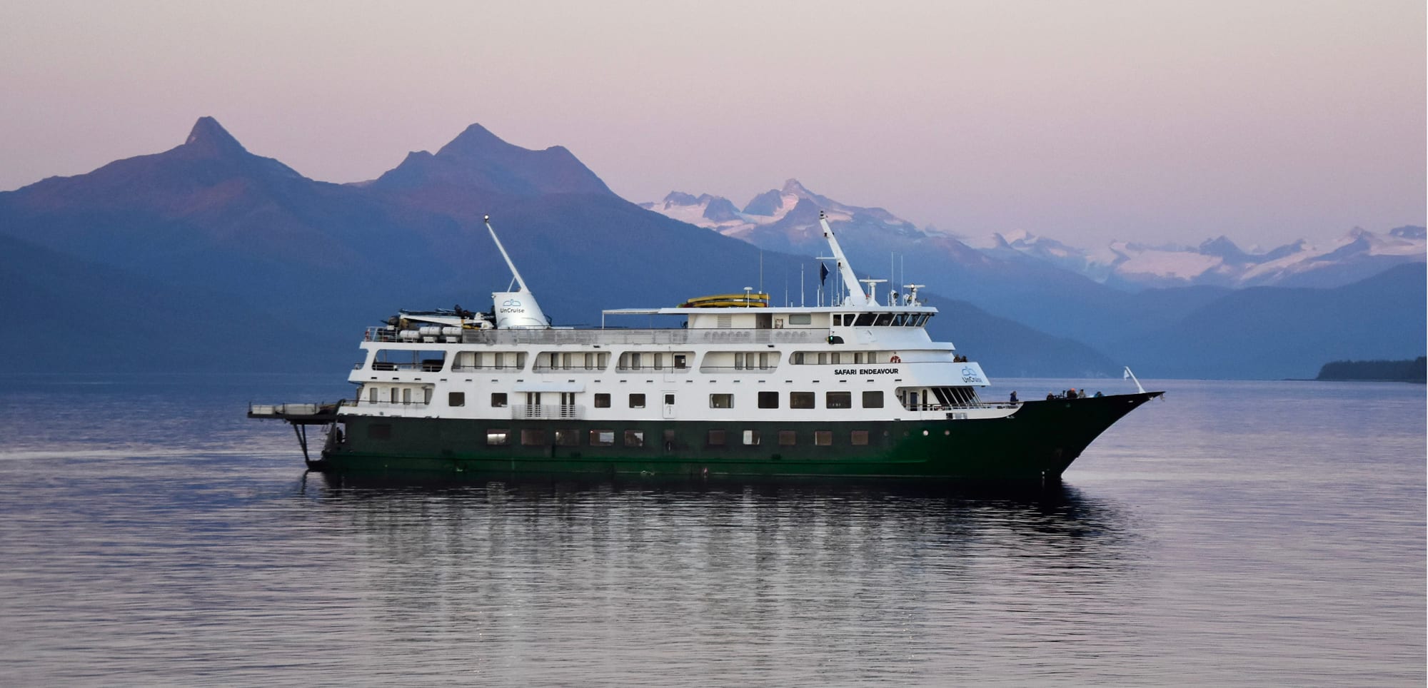 A white and green expedition cruise ship sailing on calm waters with dramatic mountain ranges and snow-capped peaks in the background during a soft-colored twilight or dawn.
