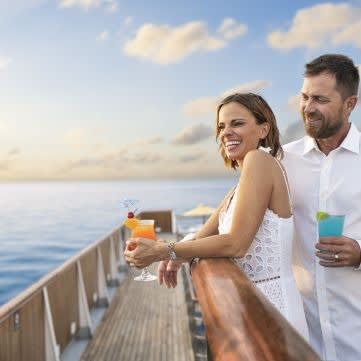 A couple stands at the railing of a cruise ship deck, holding colorful cocktails while gazing out