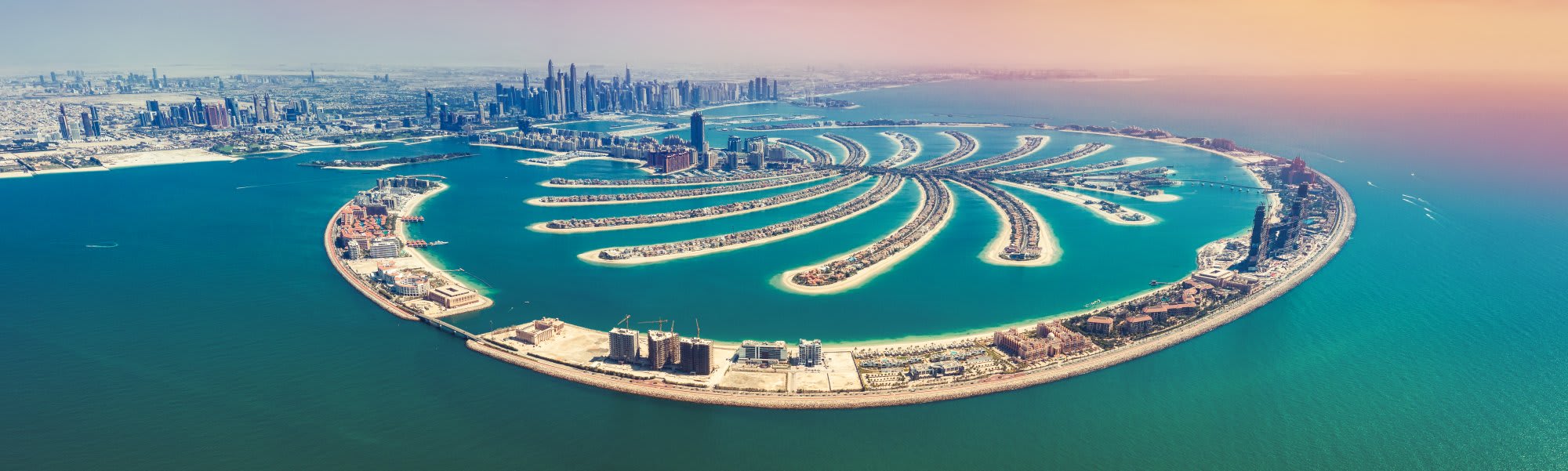 An aerial panoramic view of Palm Jumeirah, the iconic artificial palm-shaped island development in Dubai, United Arab Emirates. The image showcases the distinctive crescent-shaped waterfront with luxurious residential villas arranged in curved patterns, surrounded by turquoise Arabian Gulf waters, with the Dubai skyline and downtown cityscape visible in the background.