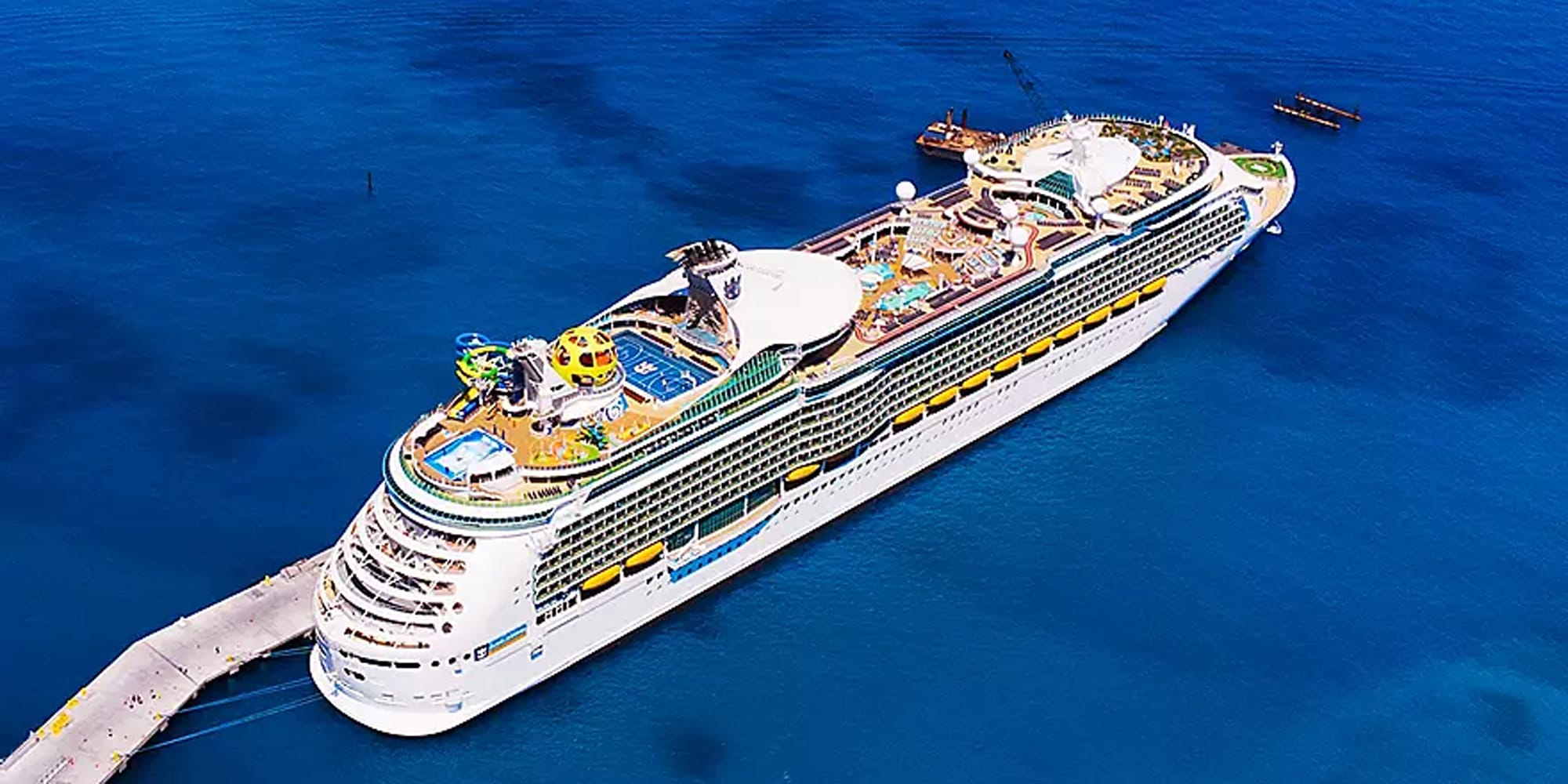 An aerial photograph of a massive modern cruise ship docked at a pier, featuring multiple decks