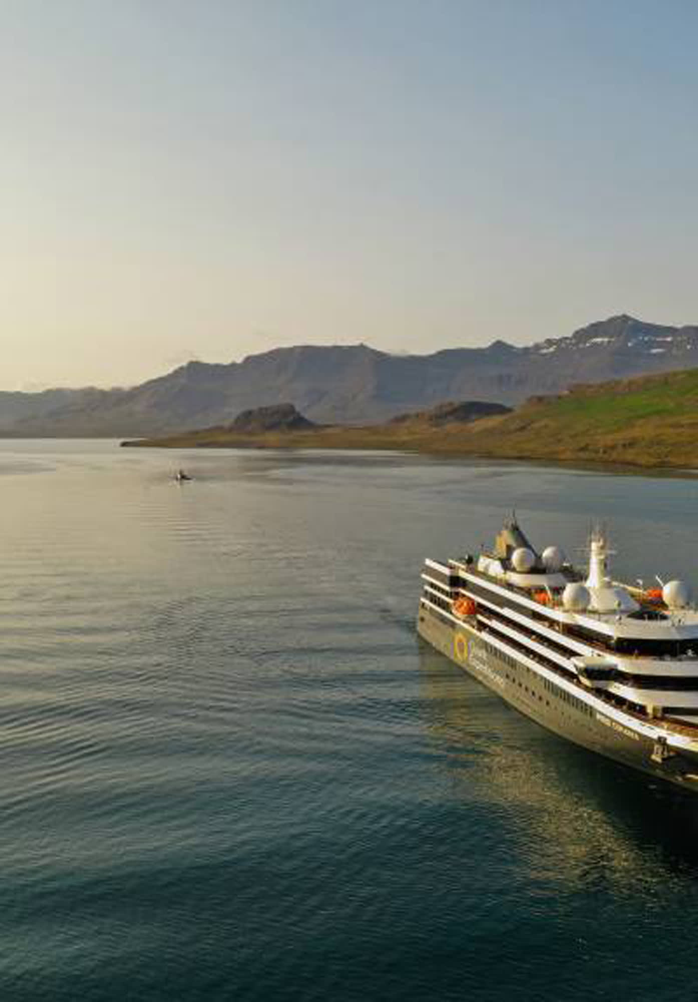 A sleek, modern cruise ship navigates through calm waters with rugged mountain terrain in
