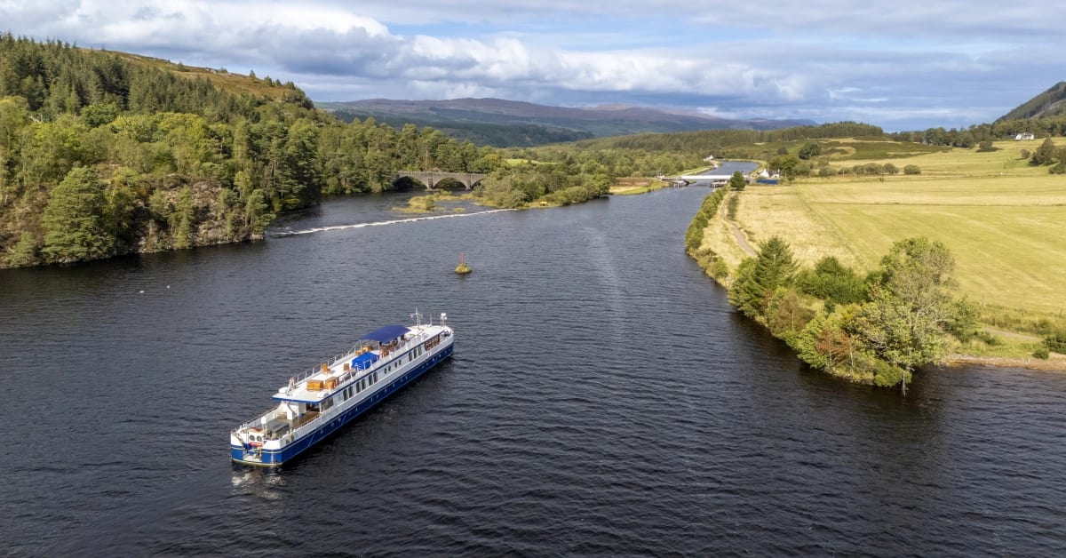 A scenic river cruise ship navigates through a picturesque Scottish valley, surrounded by lush