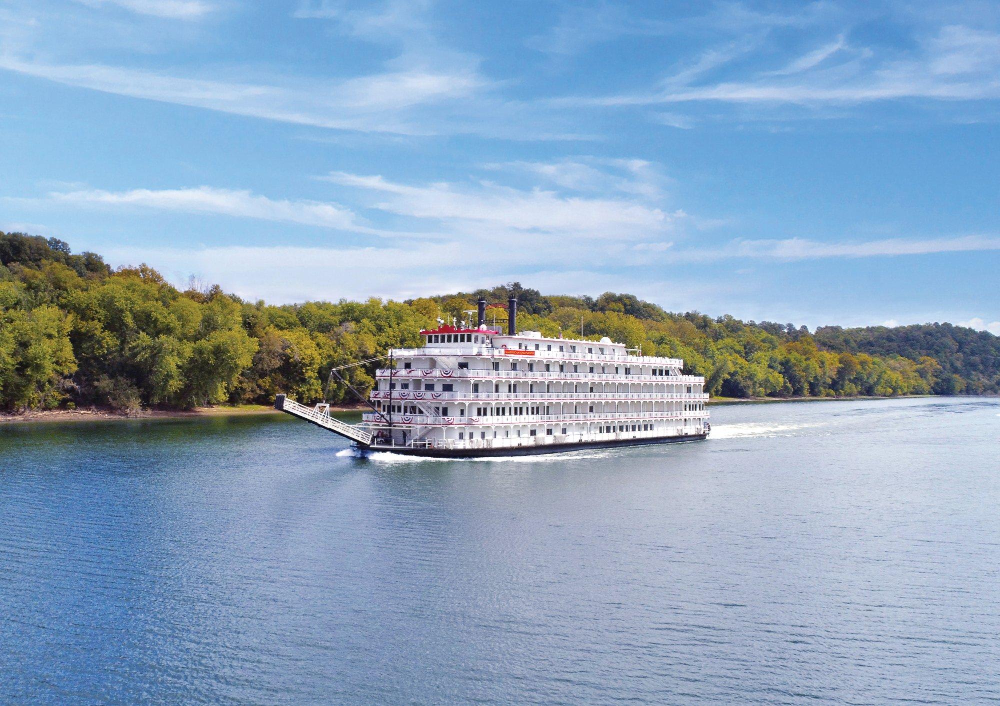 A magnificent white multi-deck paddlewheel riverboat with red trim navigates a calm river surrounded by lush green forested banks under a bright blue sky. The vessel exemplifies classic American riverboat cruising with its traditional architecture, multiple decks, and iconic paddle wheel design.