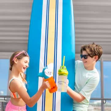 Two young children stand together in front of a large blue surfboard, exchanging colorful tropical
