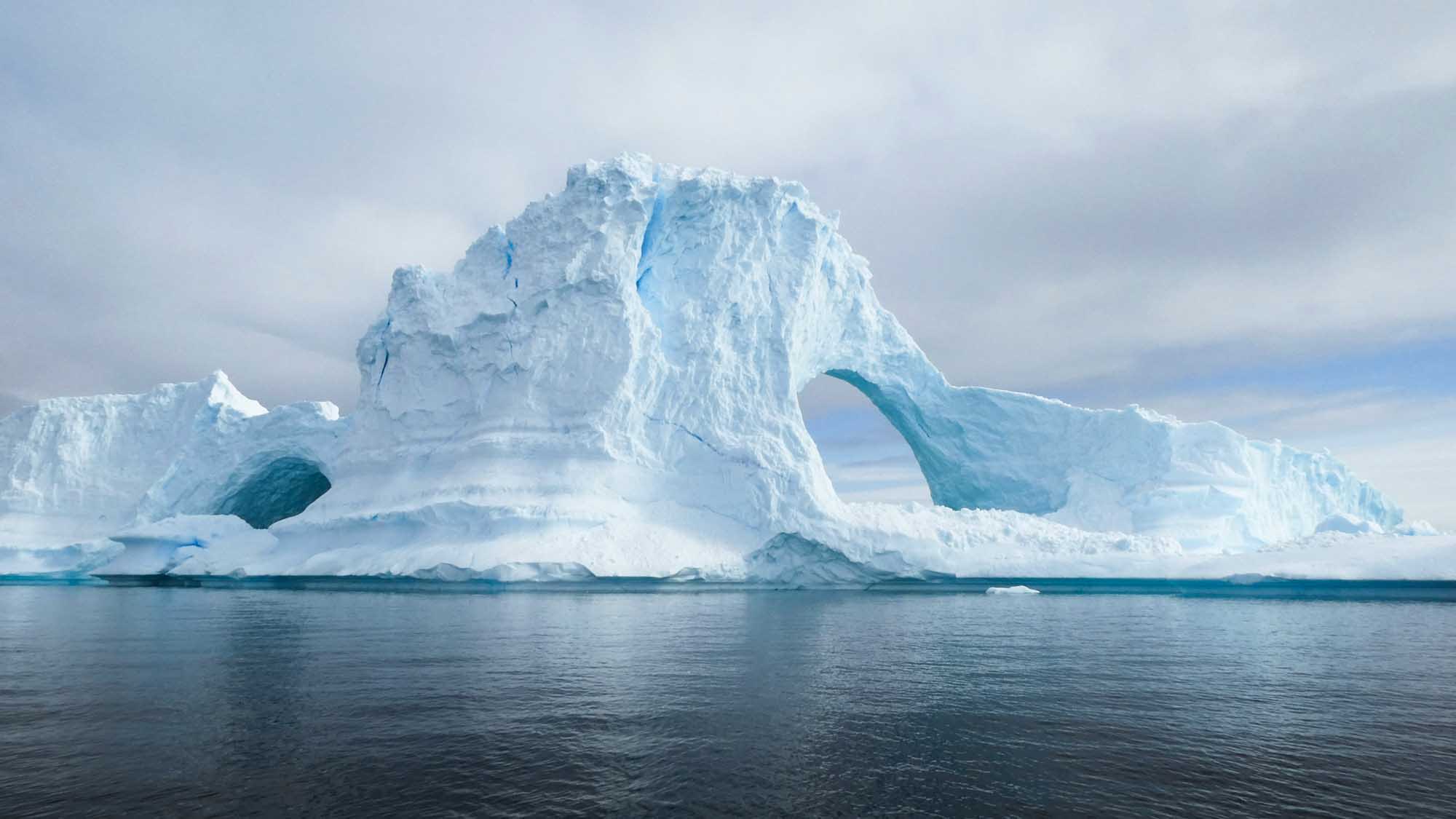 A spectacular tabular iceberg featuring a distinctive natural arch formation carved through its center, floating in calm turquoise waters. The massive ice structure displays brilliant white and blue hues characteristic of glacial ice, with its towering peak and sculpted caverns creating a striking polar landscape.