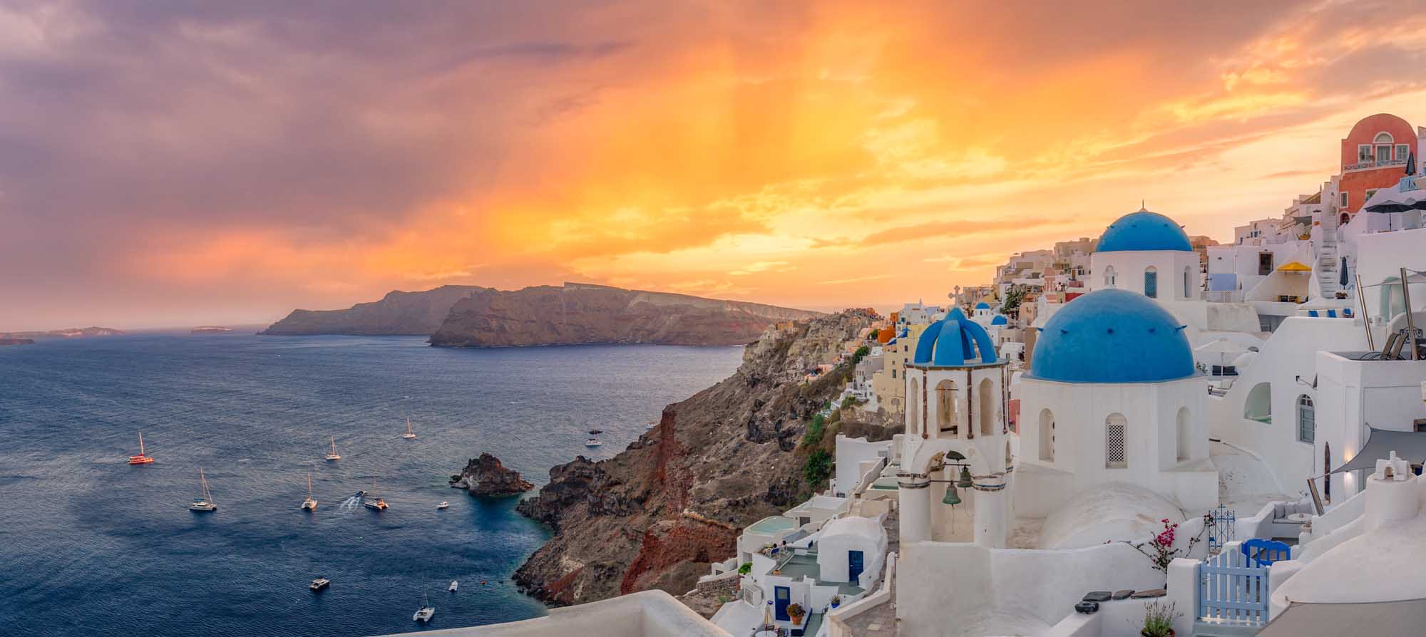 A breathtaking panoramic view of the iconic village of Oia perched on the cliffs of Santorini, Greece, featuring traditional whitewashed buildings with distinctive blue-domed churches overlooking the Aegean Sea during a stunning golden sunset. Sailboats dot the crystal-clear waters below, creating a quintessential Mediterranean luxury travel destination scene.