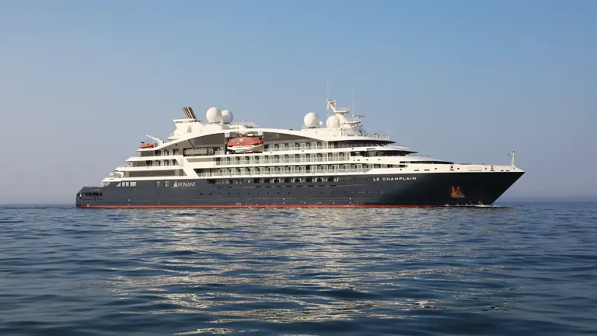 A sleek white and dark gray luxury expedition cruise ship sailing on calm blue waters. The vessel