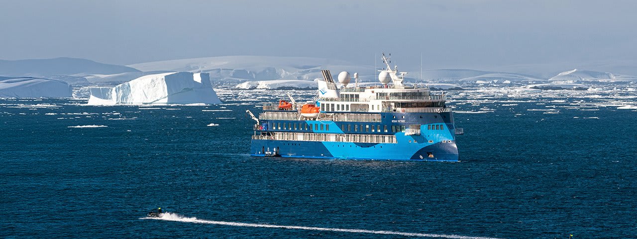 A modern blue and white expedition cruise ship navigates Arctic waters with a large tabular iceberg