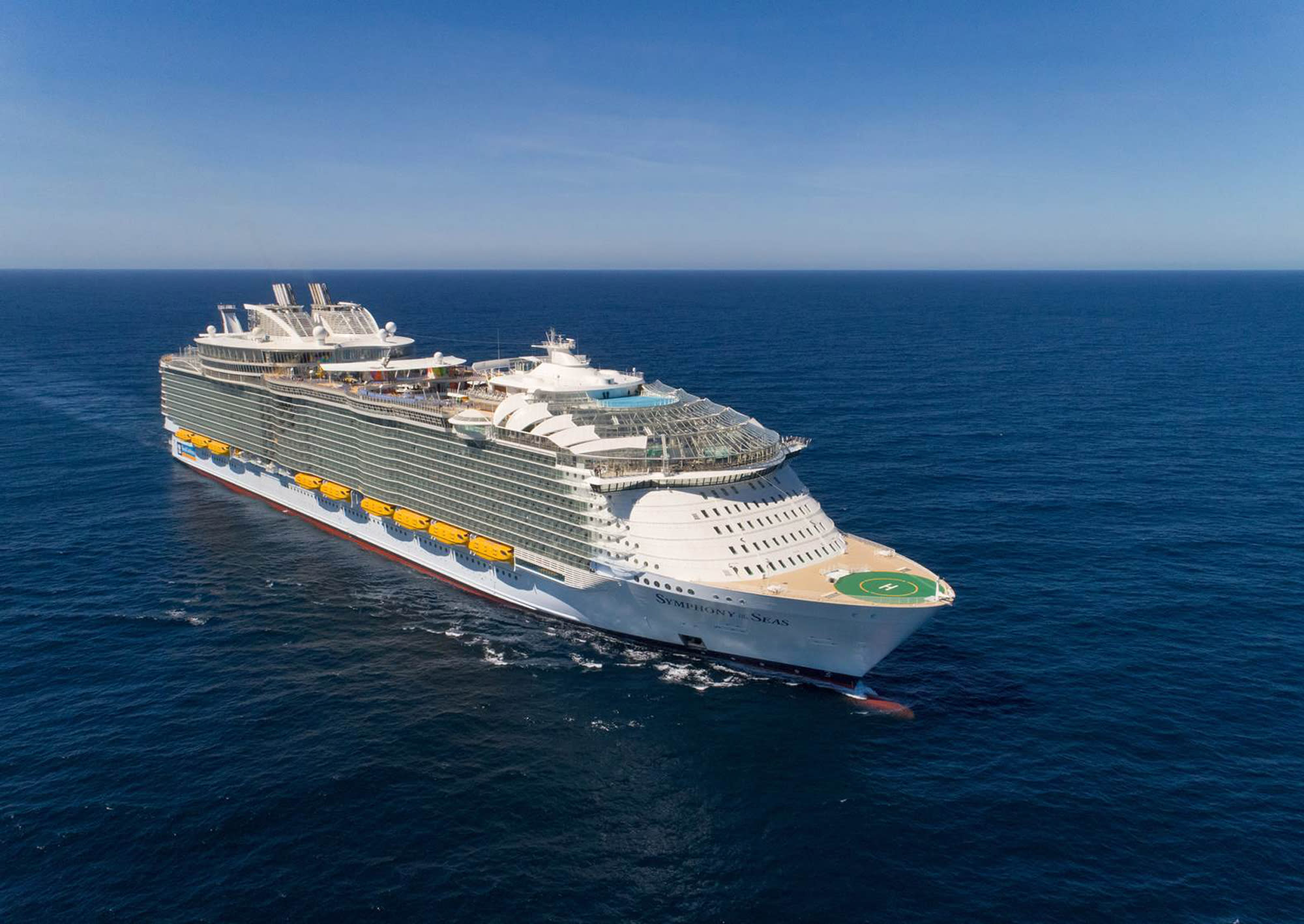 A massive modern cruise ship photographed from an aerial perspective, sailing on clear blue waters