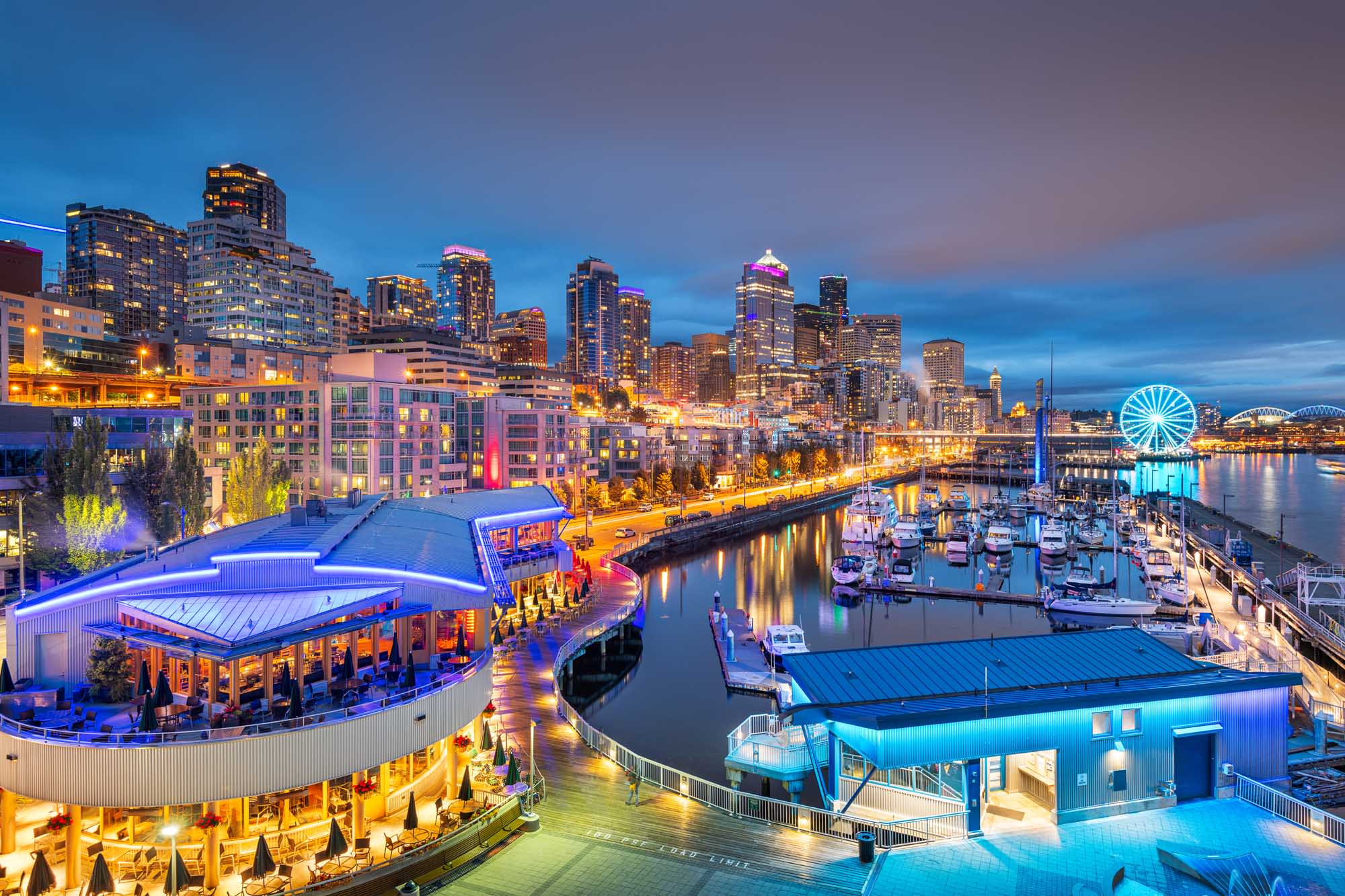A vibrant twilight view of Seattle's iconic waterfront, featuring the illuminated Great Wheel