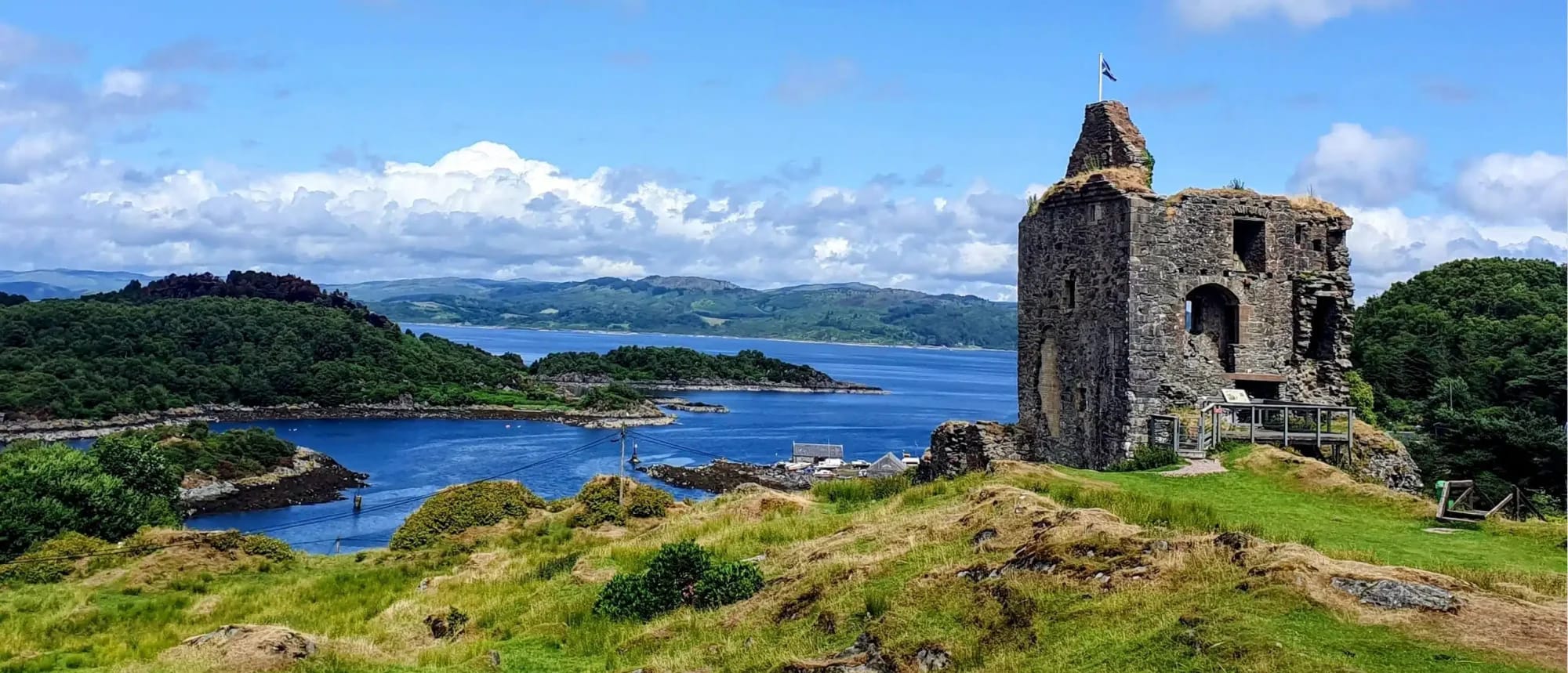 A dramatic stone castle ruin with a flagpole stands on a green clifftop overlooking a picturesque