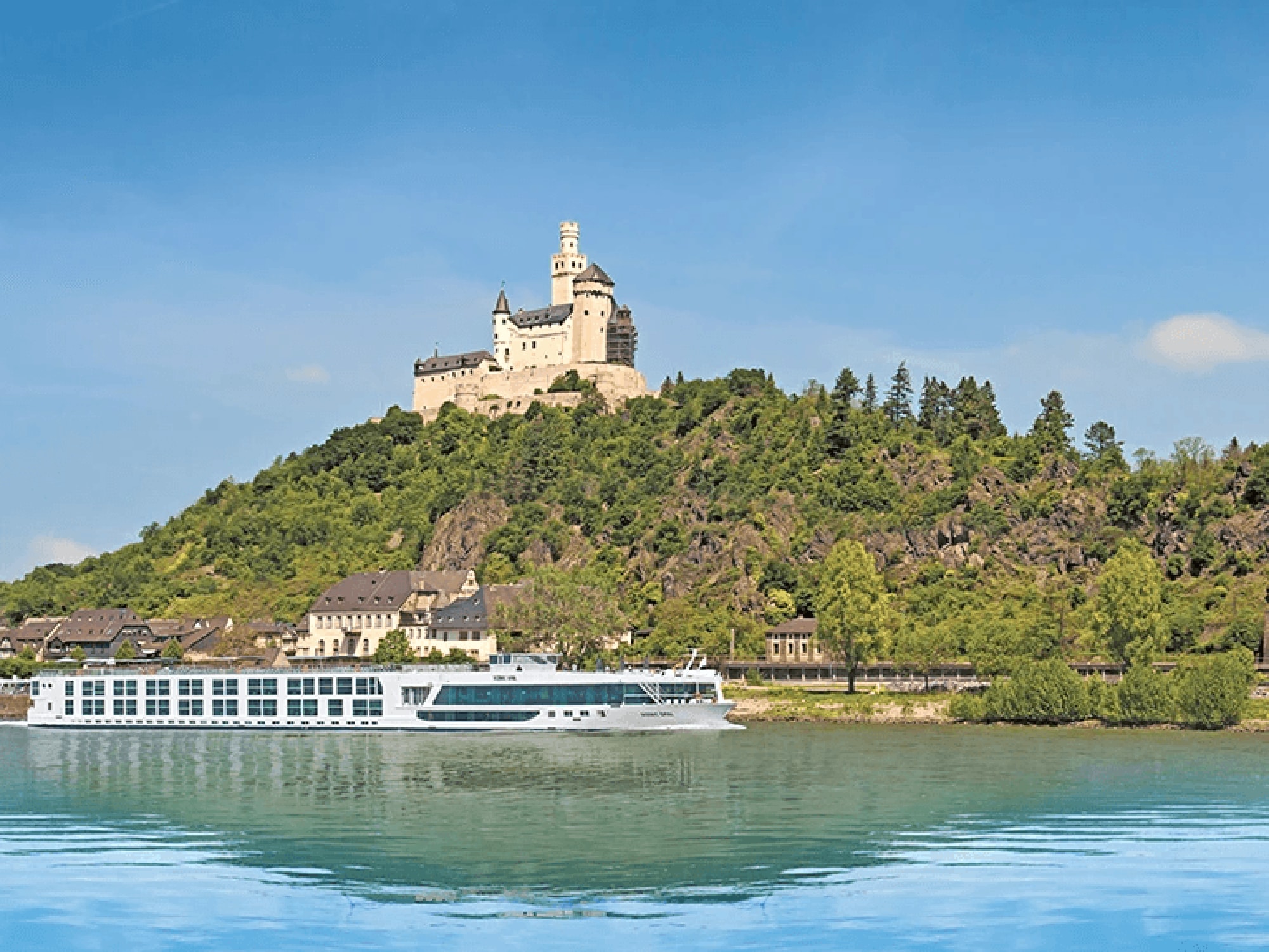 A majestic medieval castle perched atop a forested hill overlooking the Rhine River, with a modern