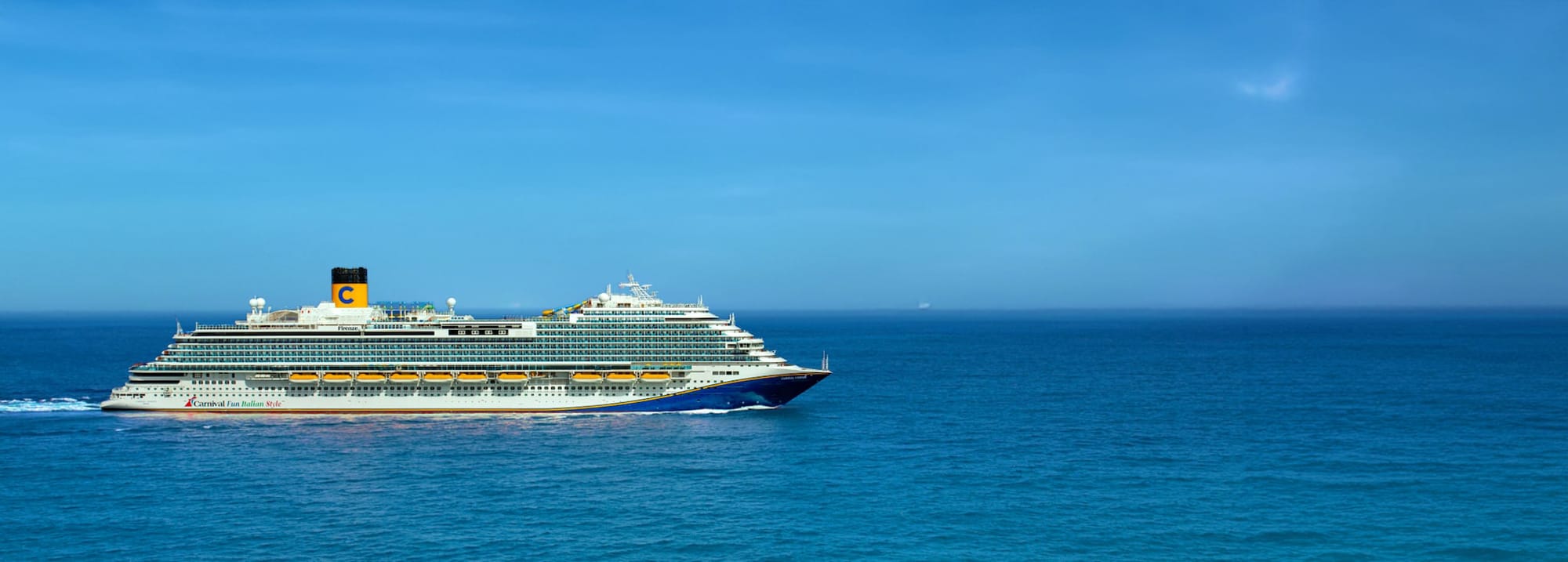 A large Carnival cruise ship sailing on a calm, vibrant blue ocean under a clear sky. The vessel
