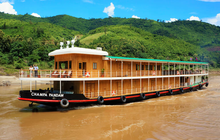 A wooden river cruise boat named Champa Pandaw sailing on a muddy river with green mountainous terrain in the background. The vessel appears to be a traditional style river cruiser with multiple decks and wooden construction.
