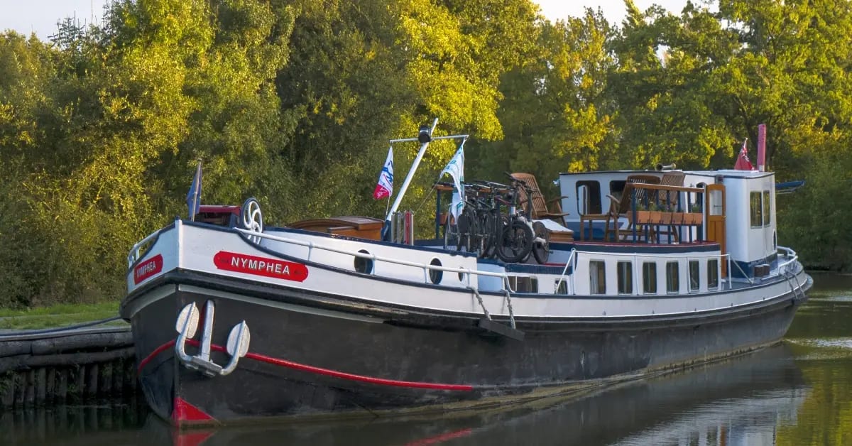 A charming traditional European barge named Nymphea is moored along a peaceful riverside, featuring