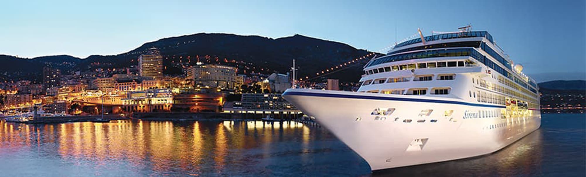 A large white cruise ship moored at a vibrant nighttime port with illuminated city buildings