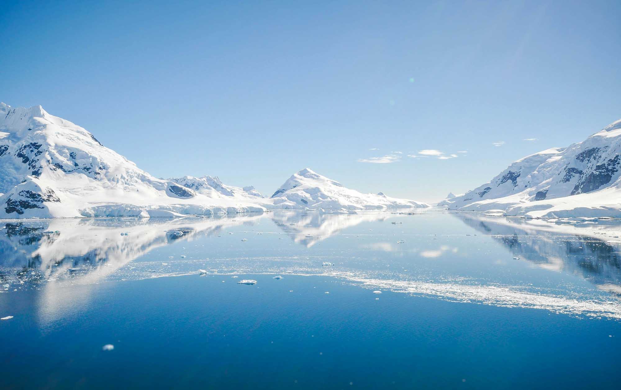 A breathtaking Arctic or Antarctic landscape featuring pristine snow-covered mountains reflected in calm, clear waters dotted with icebergs. The serene scene captures the majesty of polar regions with towering peaks and glacial ice under a bright, clear blue sky.