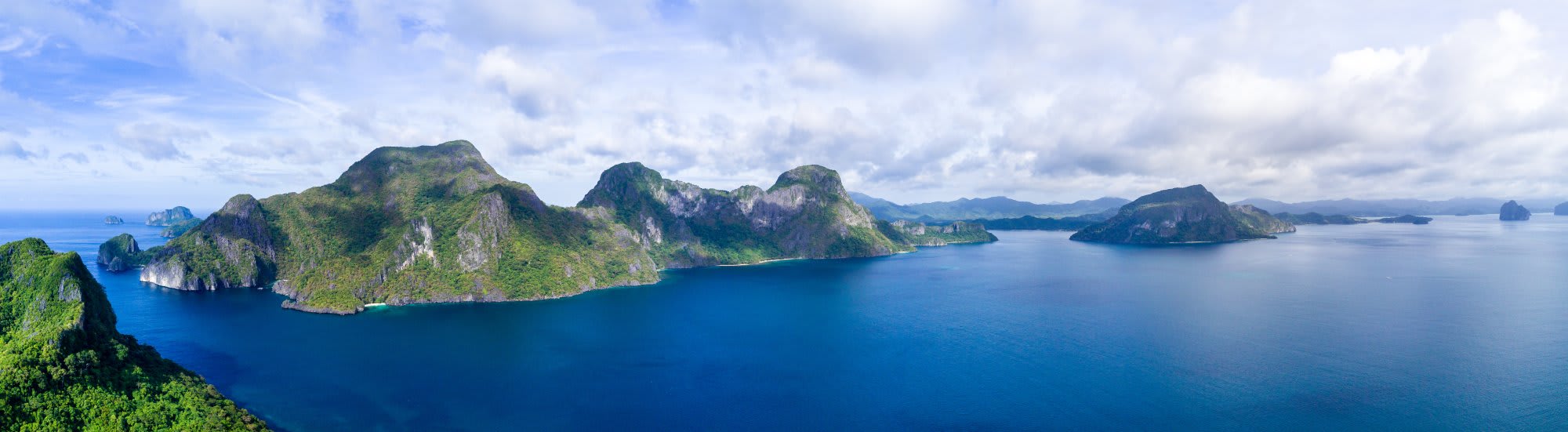 A stunning aerial view of a lush tropical archipelago featuring steep limestone cliffs covered in dense green vegetation, rising dramatically from crystal-clear turquoise waters. Multiple islands of varying sizes are scattered across the seascape under a partly cloudy sky, suggesting a Southeast Asian destination popular with cruise itineraries.