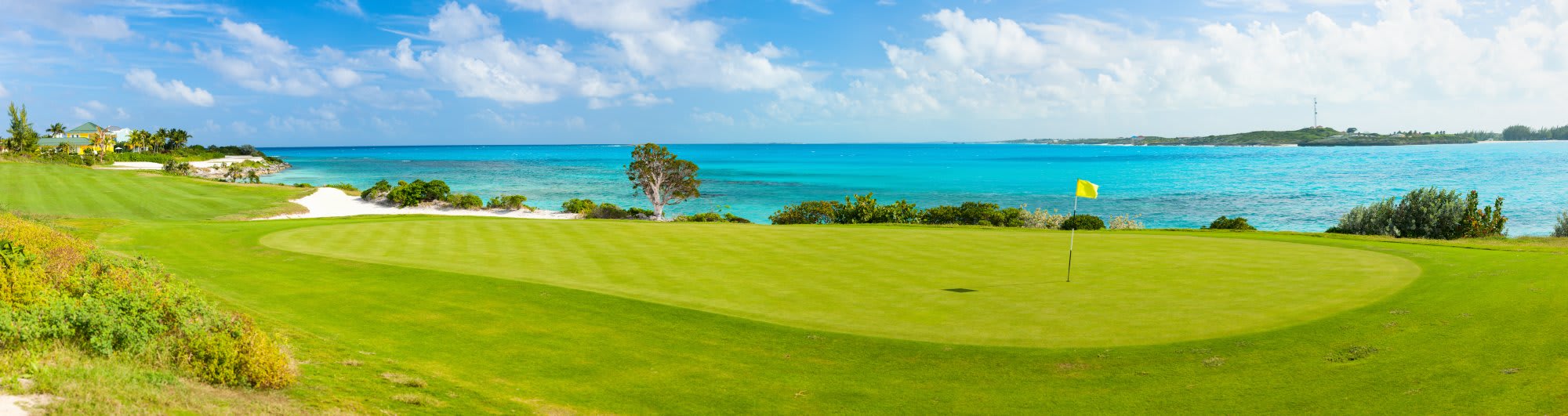 A scenic coastal golf course featuring a pristine green fairway overlooking crystal-clear turquoise waters and a white sand beach, with lush tropical vegetation and a yellow flag marking the pin in the distance.