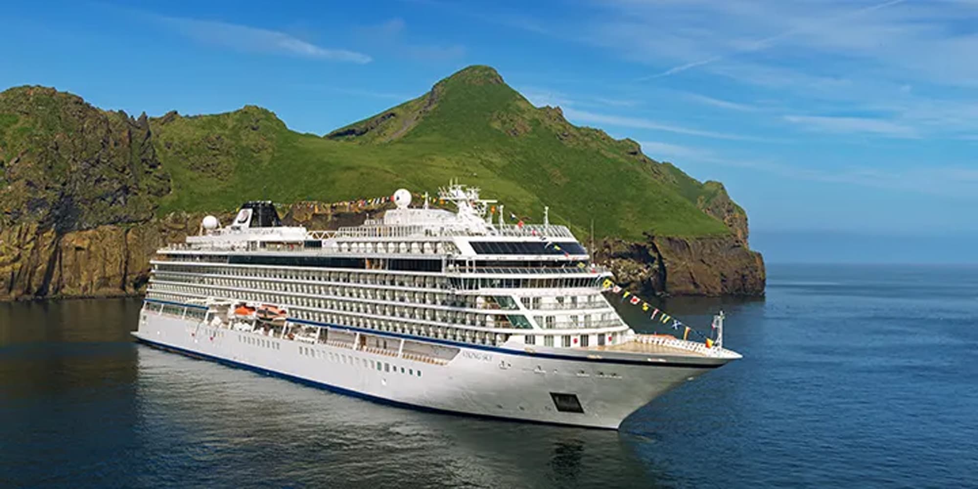 A white luxury cruise ship sailing near a green mountainous island with rocky cliffs and a calm blue ocean. The vessel is adorned with colorful flags and positioned against a picturesque volcanic landscape.