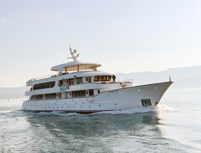 A stunning white multi-deck luxury yacht anchored in crystal-clear turquoise Mediterranean waters under a bright blue sky. The vessel features elegant modern architecture with multiple passenger decks and appears to be a premium cruise or charter yacht positioned in a pristine tropical or subtropical coastal location.