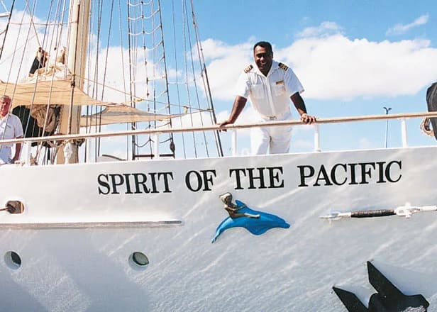 A crew member in white uniform stands on the deck of the Spirit of the Pacific, a traditional tall