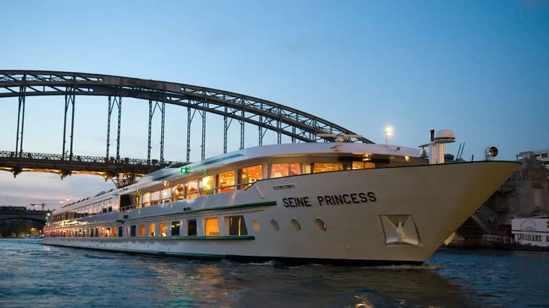 The Seine Princess, an elegant river cruise ship, is illuminated at dusk as it passes beneath
