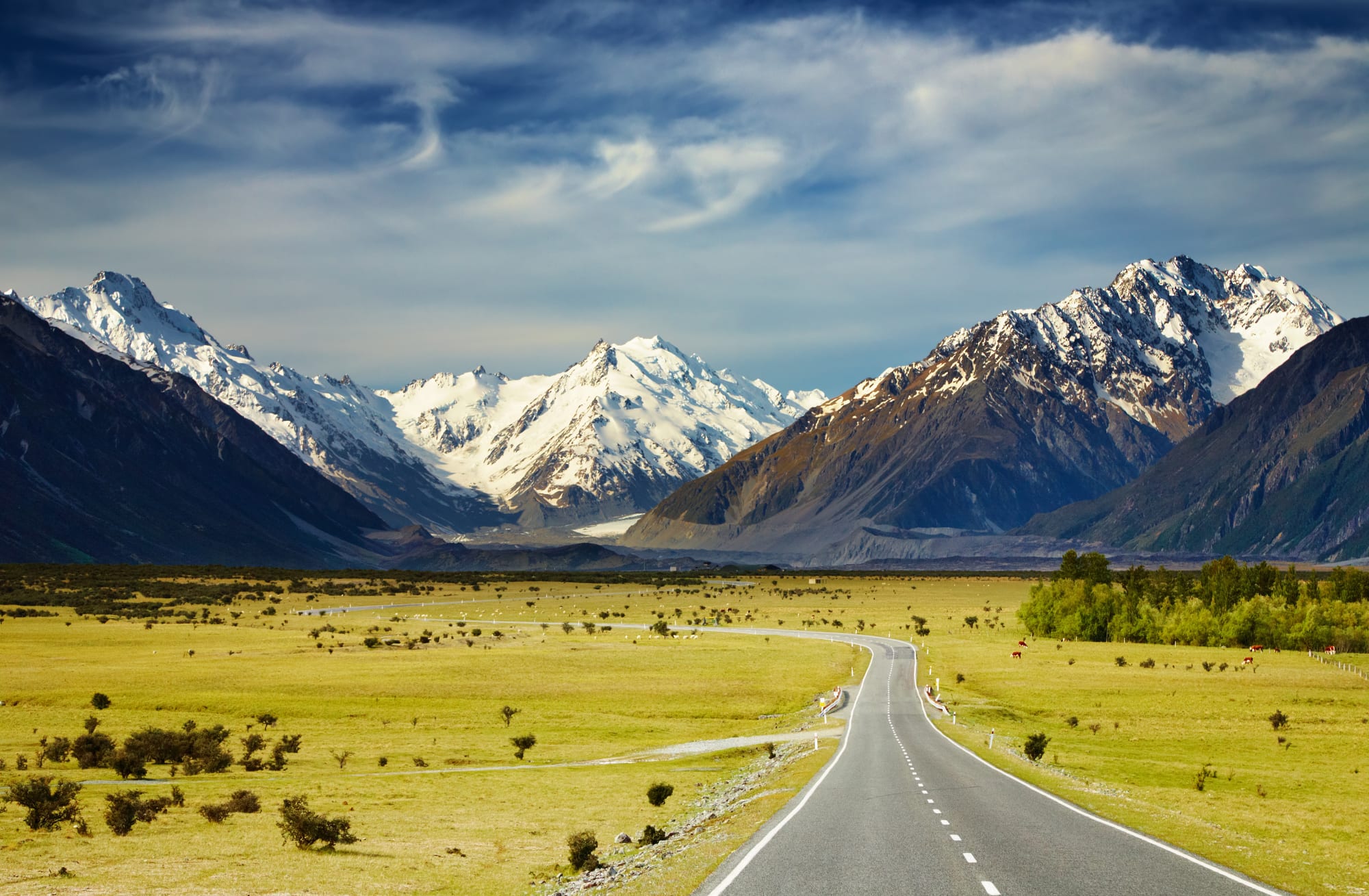 A straight asphalt highway stretches through a verdant alpine valley dotted with sparse vegetation