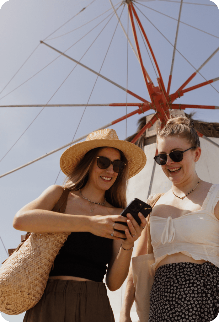 Two smiling women on a cruise ship deck, wearing sunglasses and summer attire, looking at a mobile