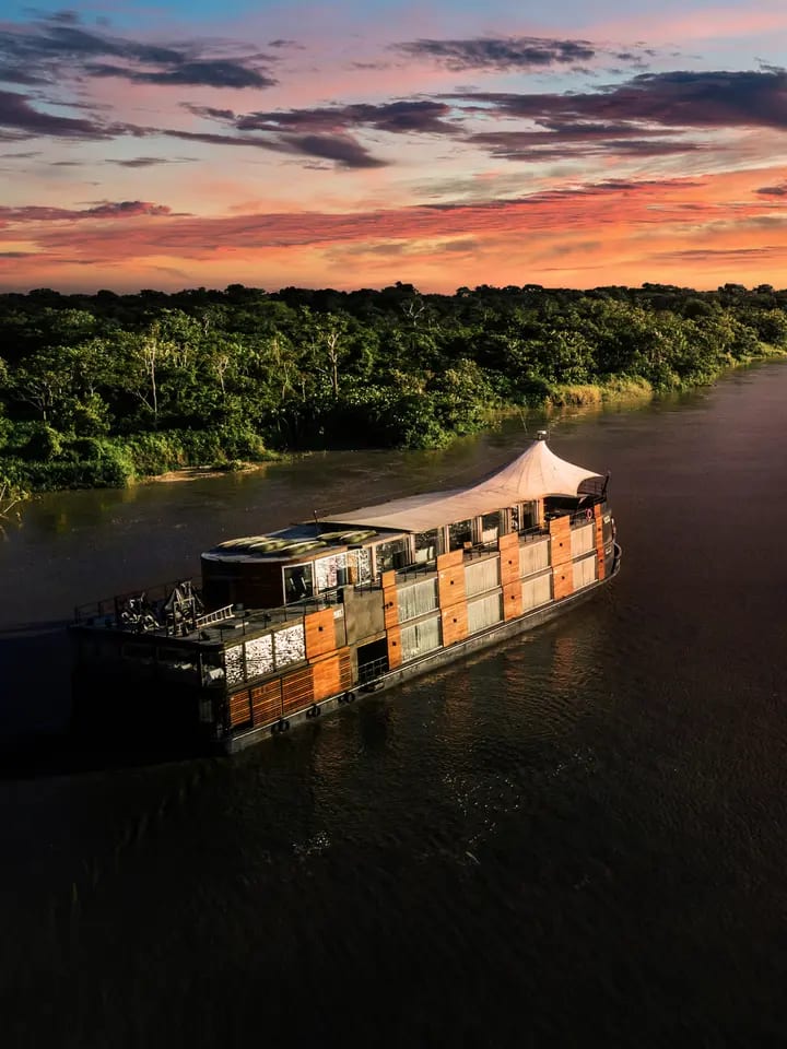 A traditional wooden riverboat cruise vessel navigates the Amazon River during a stunning golden