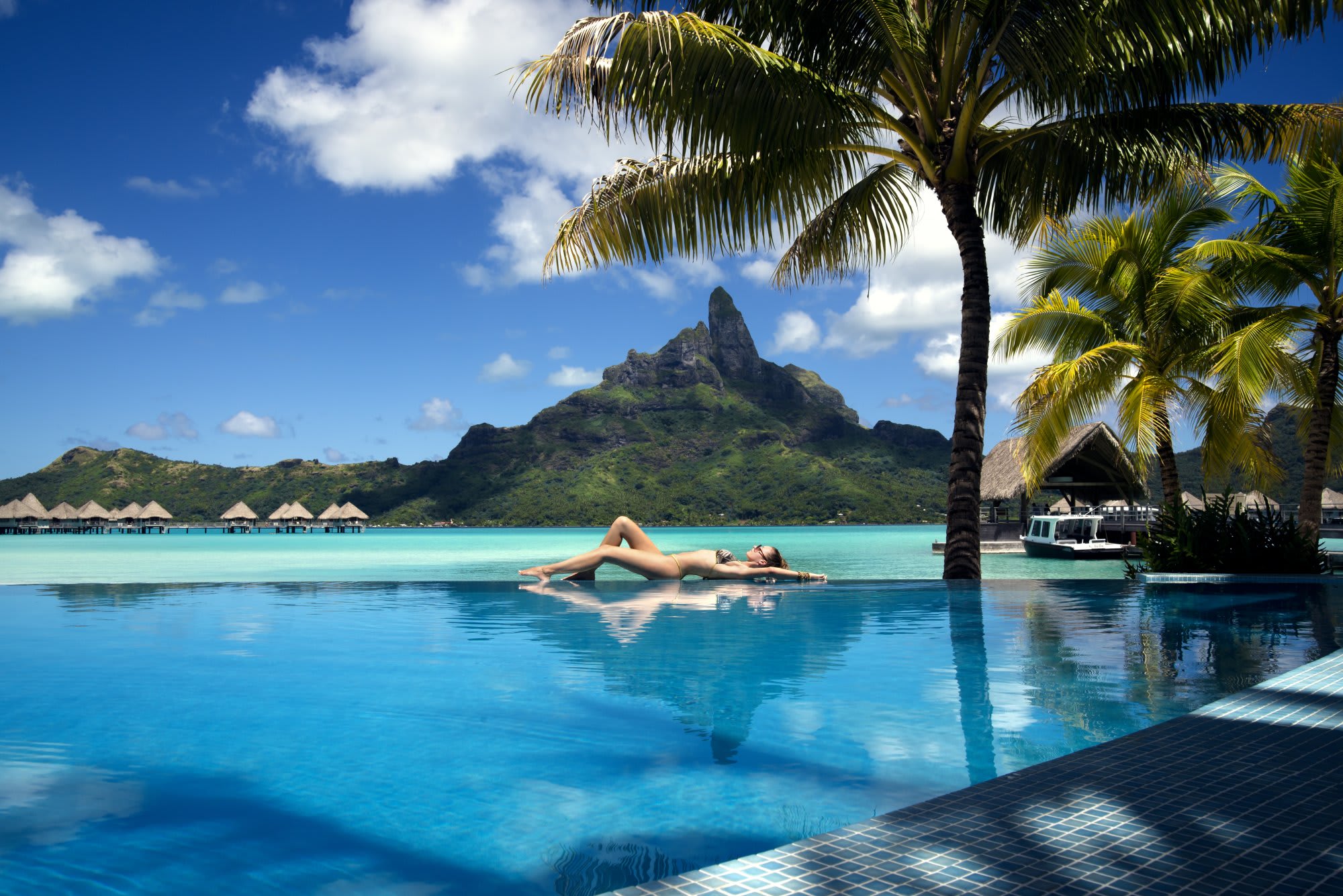 A woman relaxes on a floating lounger in an infinity pool at a tropical resort, with crystal-clear turquoise water, palm trees, and a dramatic volcanic peak in the background. The serene scene captures the essence of a luxurious South Pacific island getaway with overwater bungalows visible along the shoreline.