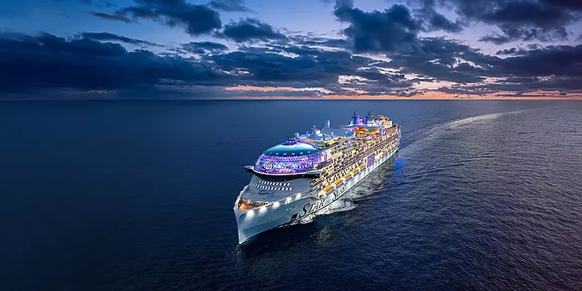 An aerial photograph of a large modern cruise ship illuminated against a dramatic twilight sky