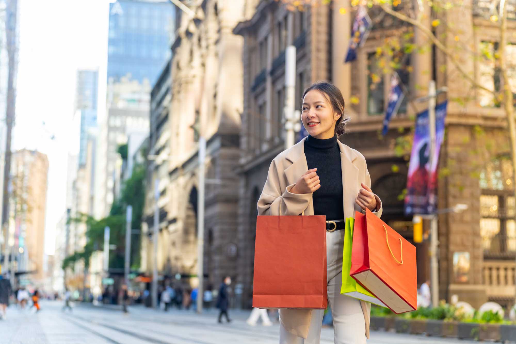 A stylish Asian woman carries colorful shopping bags while walking through a vibrant city street