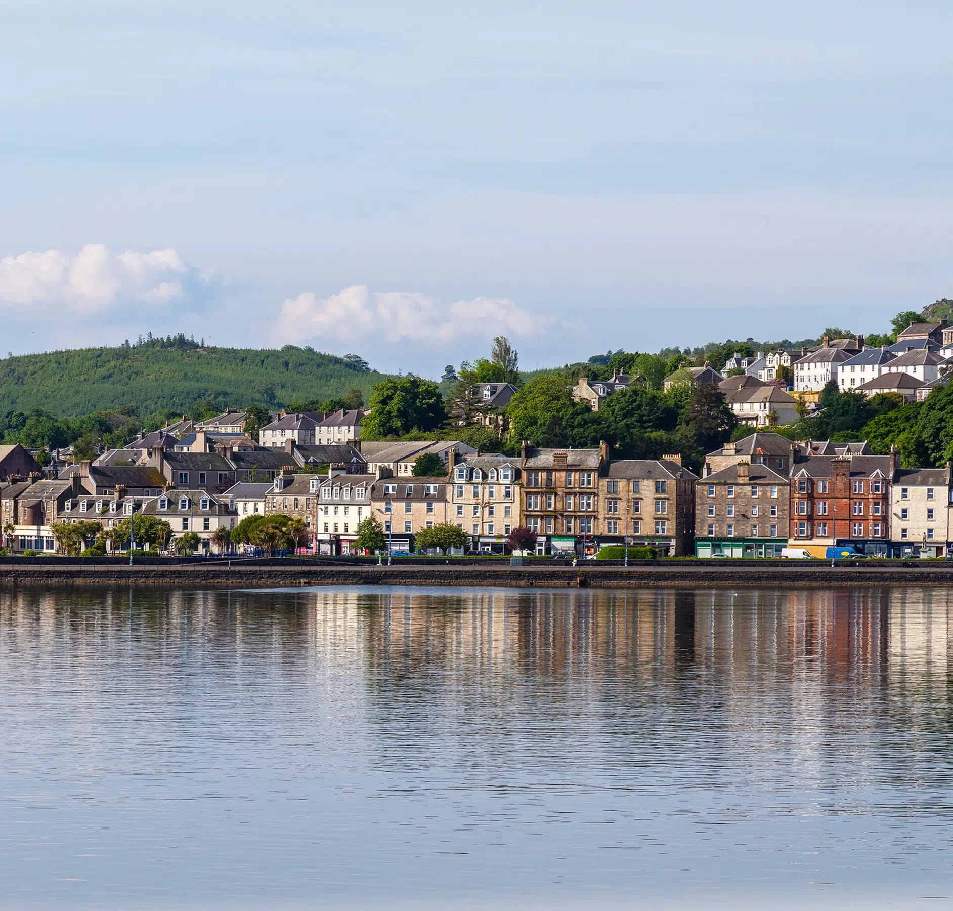 A picturesque Scottish riverside town featuring a row of traditional Georgian and Victorian stone buildings reflected in calm waters, with forested hills rising in the background. The charming waterfront townscape with its varied architecture and natural setting exemplifies classic Scottish Highlands destinations popular with cruise itineraries.