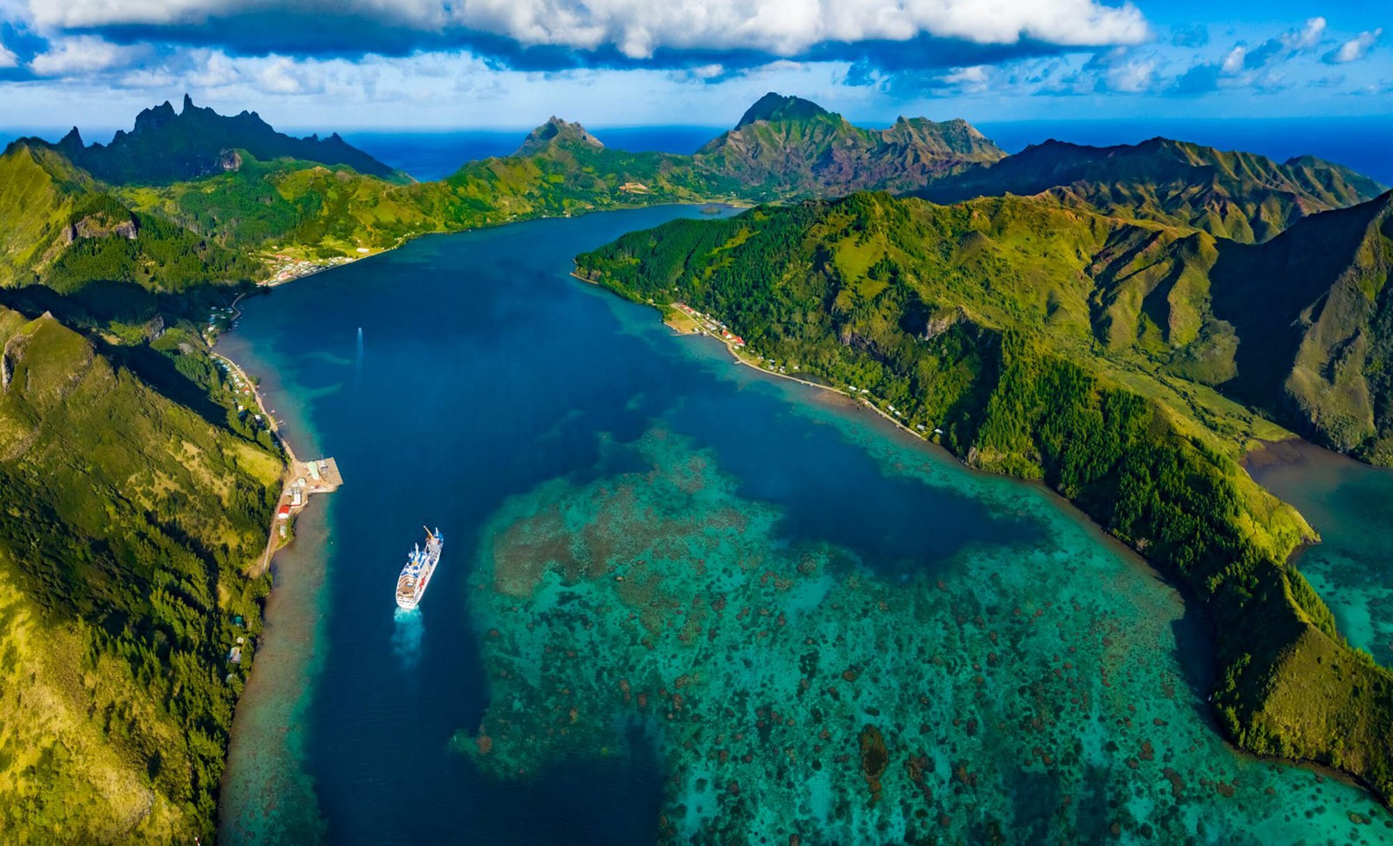 An aerial view of a cruise ship navigating through a stunning turquoise lagoon surrounded by lush green mountainous islands. The landscape features dramatic volcanic peaks and coral reefs visible beneath the crystal-clear waters.