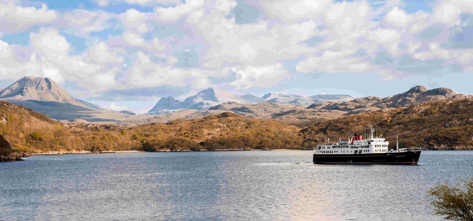 A classic expedition cruise ship anchors in calm blue waters with dramatic Scottish Highland