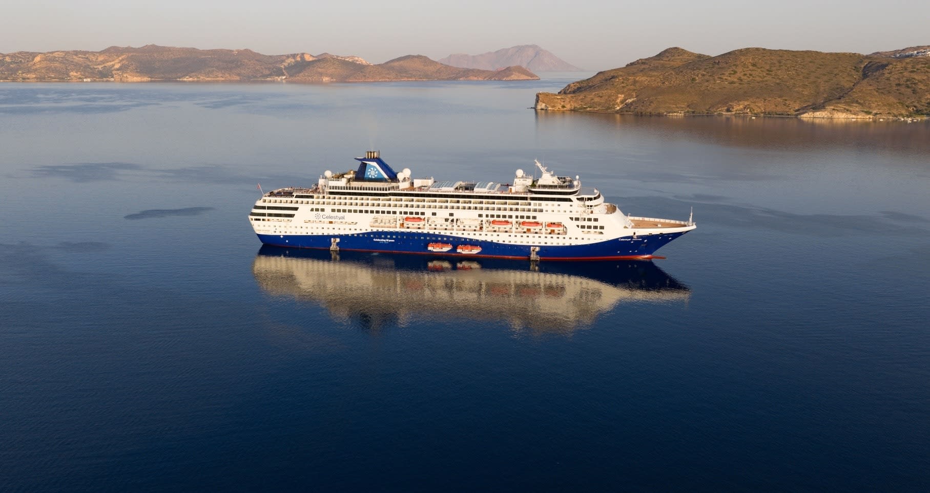 A large cruise ship operated by Celestyal Cruises anchored in calm Mediterranean waters, with arid