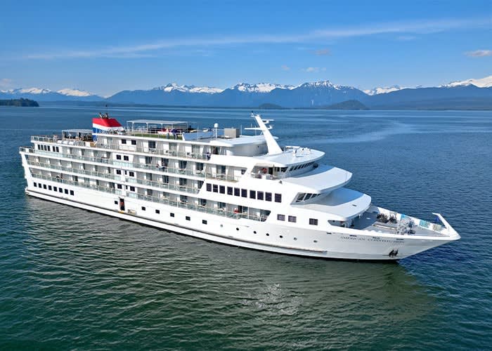 A white luxury small-ship cruise vessel navigates blue-green waters with snow-capped mountains
