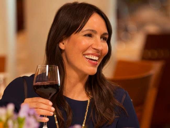 A woman smiles while holding a glass of red wine in an elegant dining room with warm lighting