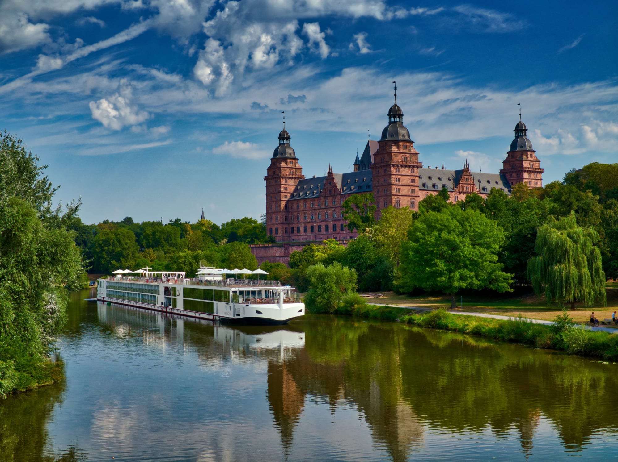 A modern river cruise ship navigates a tranquil river in front of an impressive Renaissance-style castle with multiple towers, surrounded by lush green trees and a dramatic blue sky with white clouds.