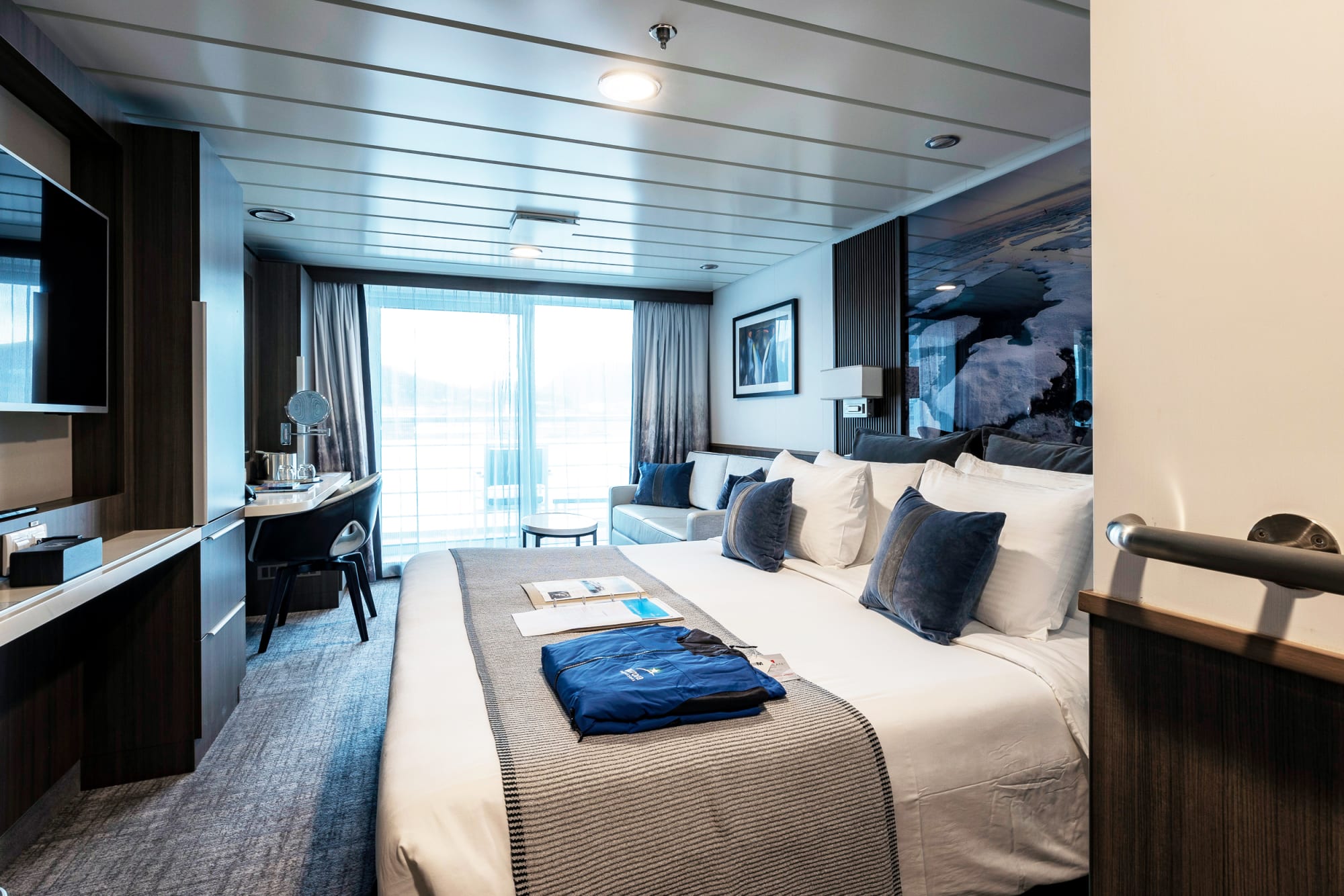 A luxurious cruise ship stateroom featuring a king-size bed with premium bedding, floor-to-ceiling windows with ocean views, modern furnishings in dark wood tones, a work desk, and sophisticated blue and white color scheme accents throughout the elegantly appointed cabin.