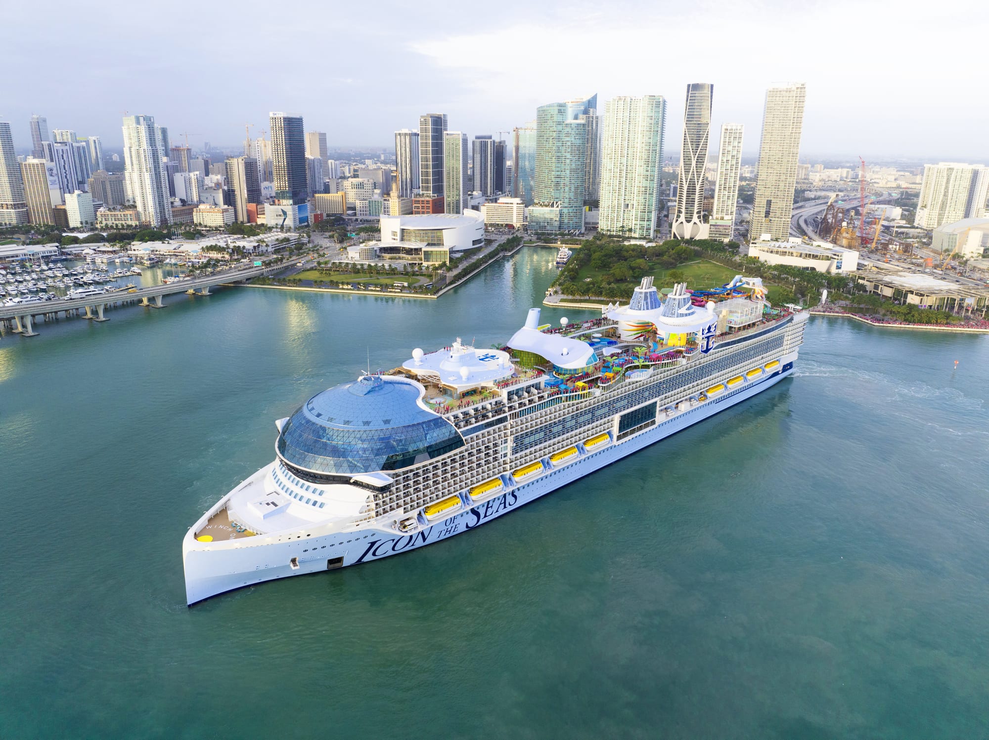 A massive modern cruise ship with distinctive white hull and blue accents is docked in turquoise