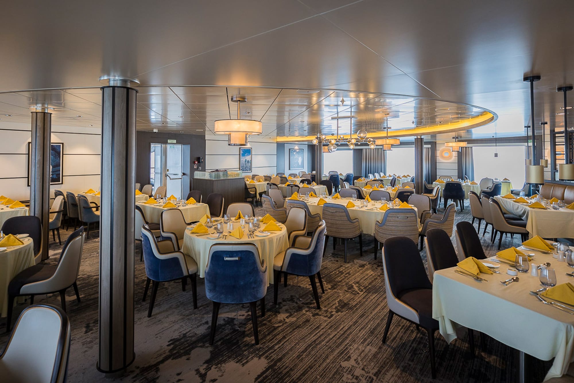 A sophisticated ship dining room featuring round tables with cream linens and yellow napkins