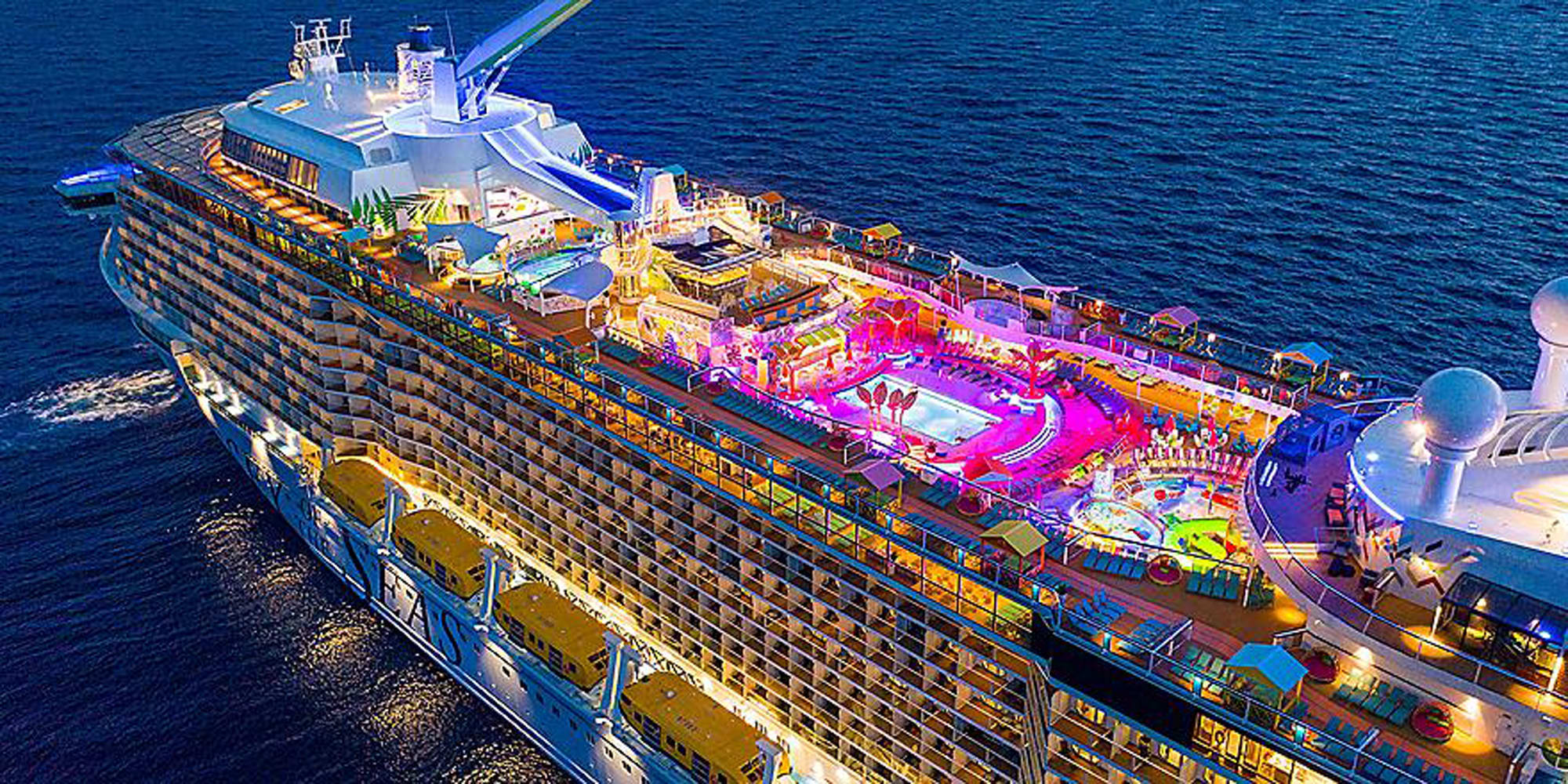 An aerial nighttime photograph of a large modern cruise ship illuminated with vibrant colored